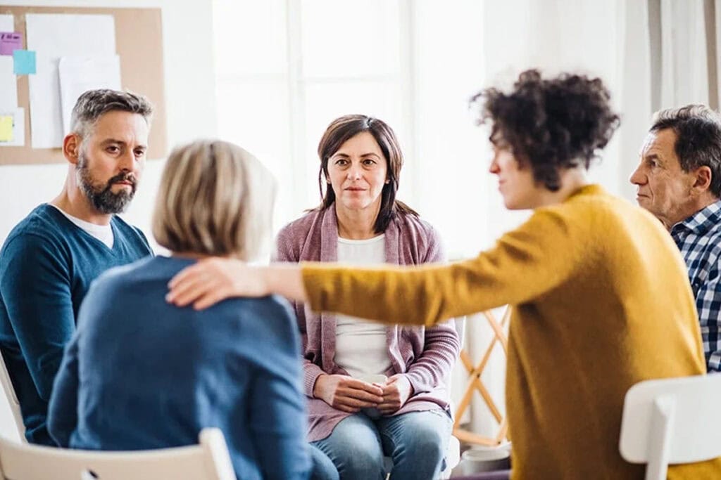 A group of five adults sit in a circle indoors, engaged in conversation. One person reaches out to comfort another, while the others listen attentively. The atmosphere appears supportive and caring.