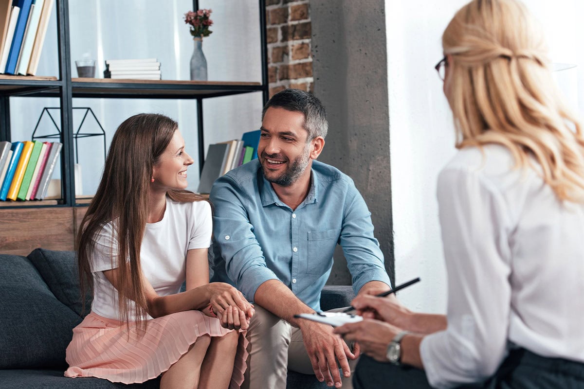 A smiling couple sits together on a couch, facing a professional woman with a notepad, suggesting a therapy or counseling session in a modern, cozy office.