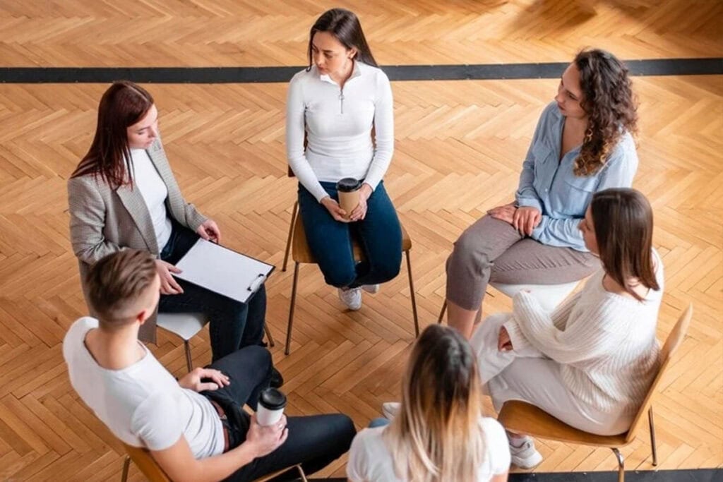 Five people sit in a circle on chairs, engaged in conversation. One person holds a clipboard, while others hold cups. They are in a room with a wooden floor, viewed from above.
