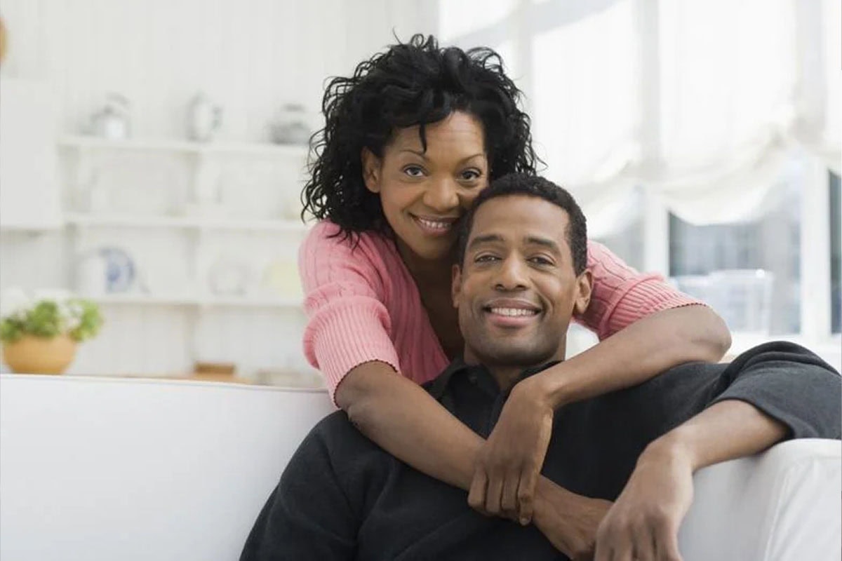 A woman in a pink sweater hugs a man in a dark sweater from behind as they smile while sitting on a white couch in a bright, cozy living room.