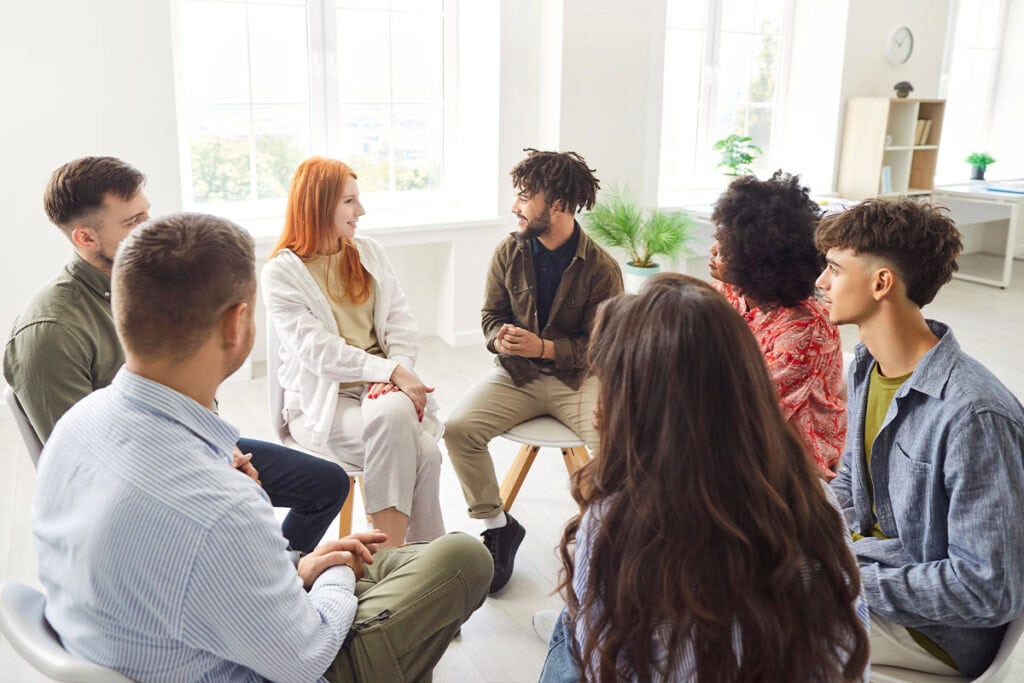 A diverse group of seven young adults sit in a circle, engaged in a discussion in a bright, modern room with large windows and natural light.