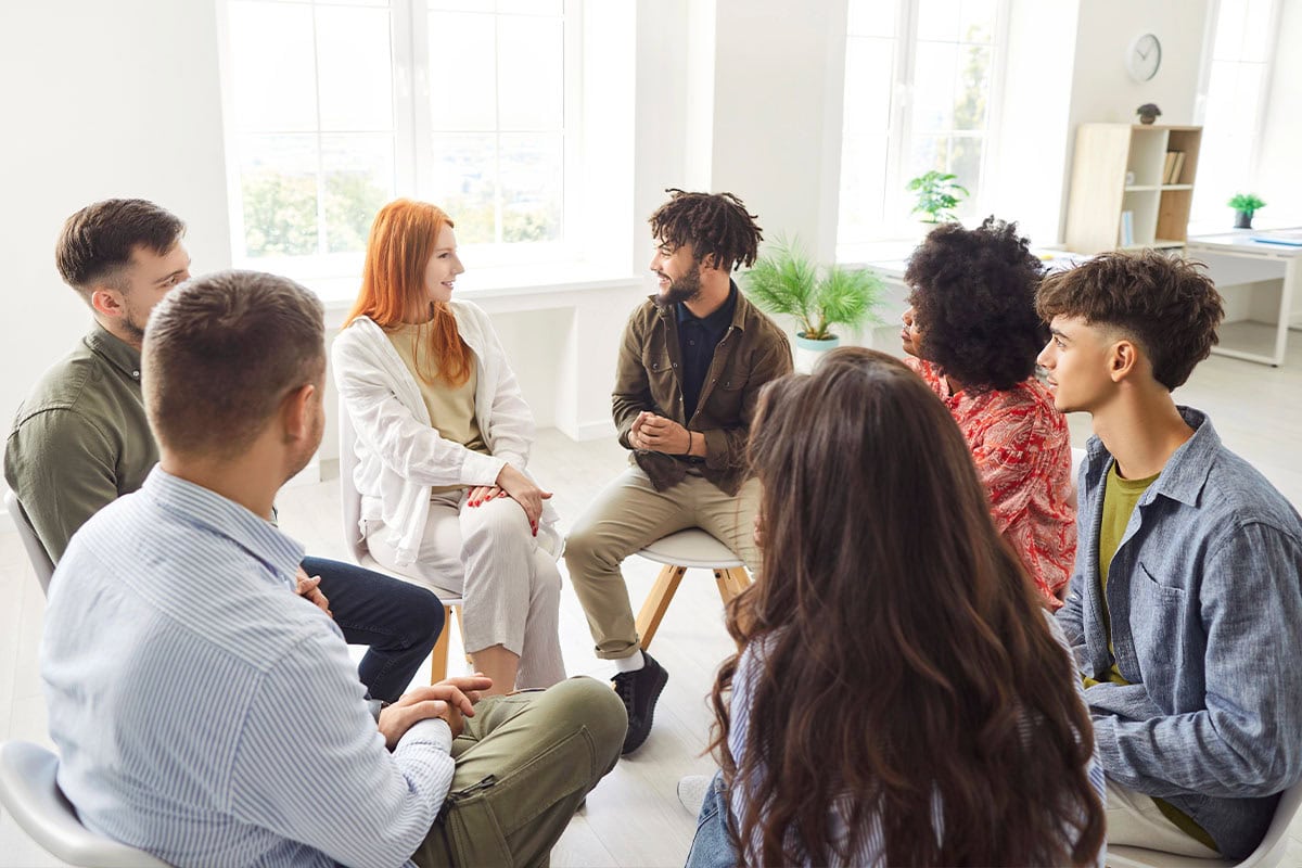 A diverse group of seven young adults sit in a circle, engaged in a discussion in a bright, modern room with large windows and natural light.
