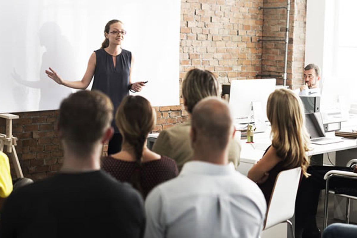 A woman stands in front of a group, giving a presentation in a modern office with exposed brick walls. Several people are seated and listening, while a man works at a computer in the background.