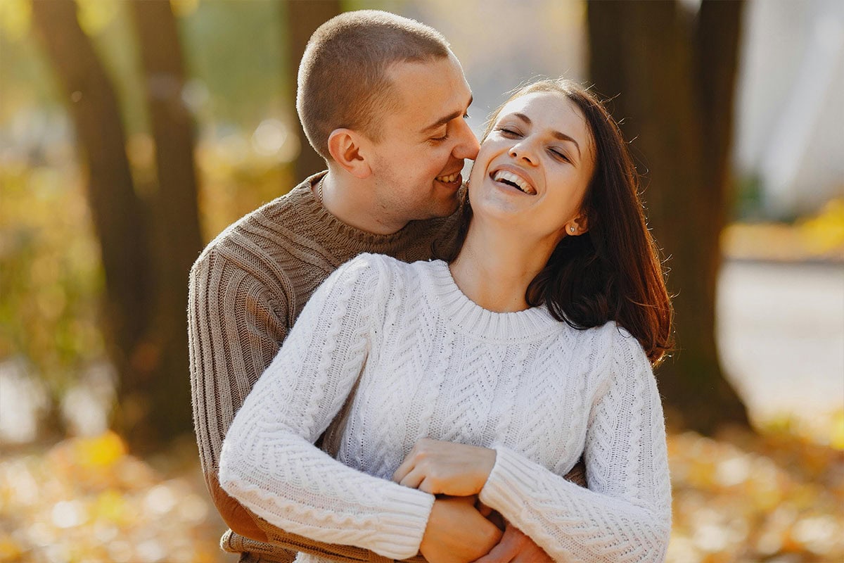 A couple is standing outdoors in an autumn park. The man hugs the woman from behind as they both smile and laugh, wearing cozy sweaters. Sunlight filters through the trees and fallen leaves cover the ground.