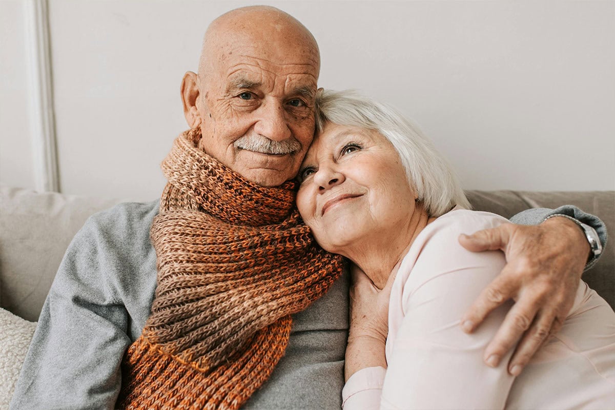 An elderly couple sits closely on a couch. The man, wearing a brown scarf, hugs the woman, who has short white hair and rests her head on his shoulder, both smiling gently and appearing content.