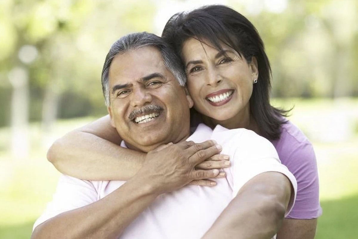 A smiling woman hugs a smiling man from behind while they sit outdoors in a park, both wearing casual shirts and looking happy.