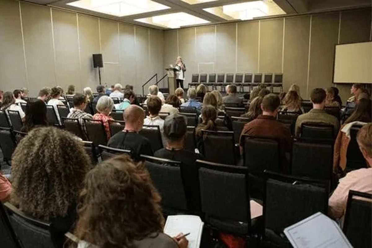 A speaker stands at a podium in front of an audience seated in rows in a conference room. The room has beige walls, a projector screen, and a row of empty chairs on stage. People are listening and taking notes.