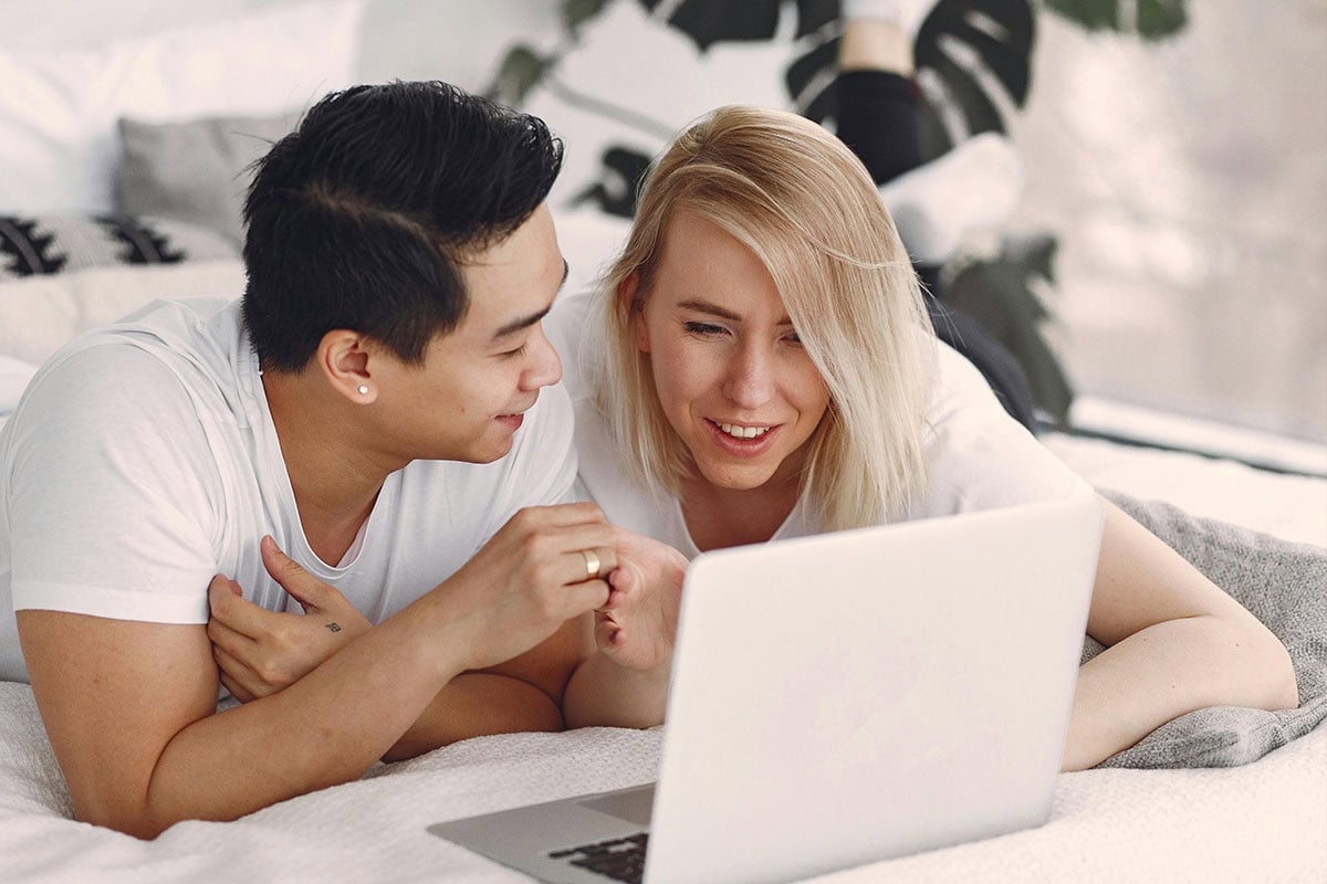 A young couple lies on a bed, smiling and looking at a laptop screen together. They appear relaxed and happy, both wearing white shirts in a bright, cozy room.
