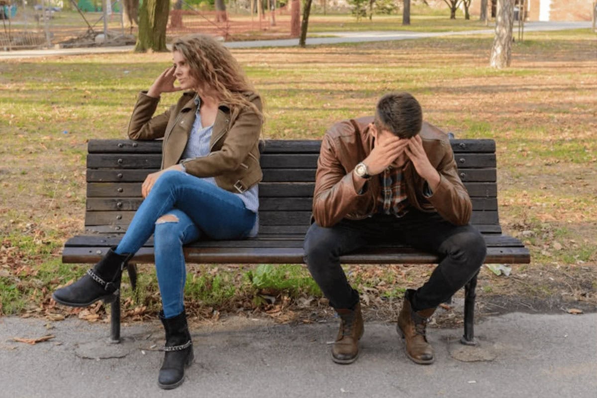 A woman and a man sit on a park bench. The woman looks away with a frustrated expression, while the man sits with his head in his hands, suggesting they are upset or having an argument.