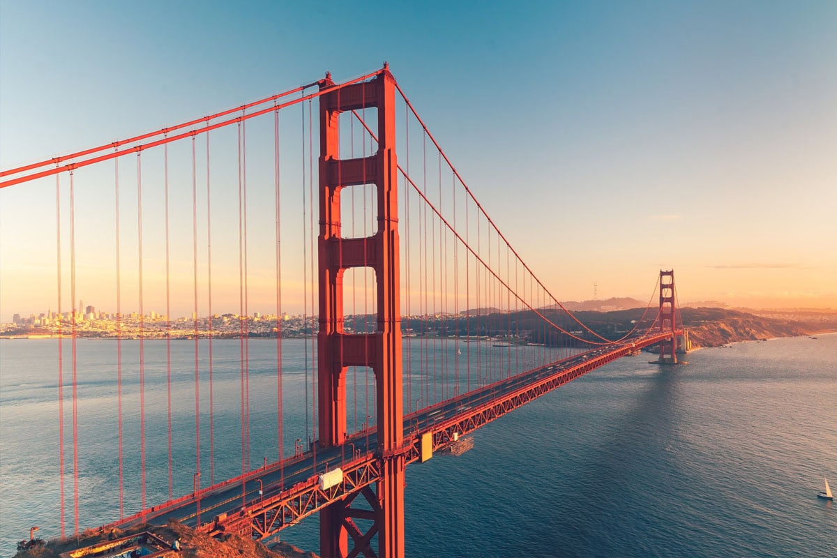 The Golden Gate Bridge stretches across blue water at sunset, with the city skyline visible in the distance and warm sunlight illuminating the iconic red suspension structure.