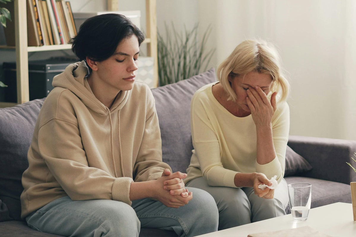 Two women sit on a couch in a living room. One looks down with clasped hands, while the other covers her face with one hand and holds a tissue, appearing upset. A glass of water is on the table in front of them.
