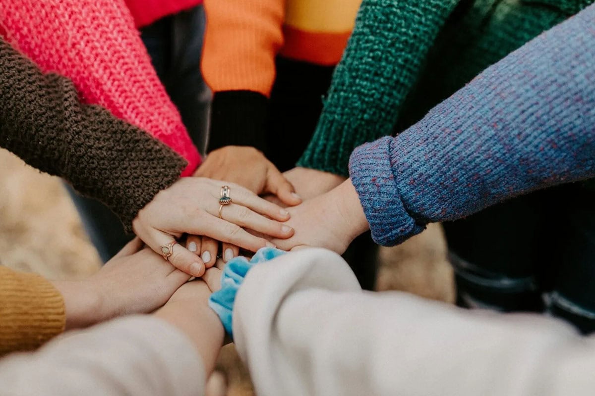 A group of people wearing colorful sweaters join their hands together in the center, symbolizing unity and teamwork.