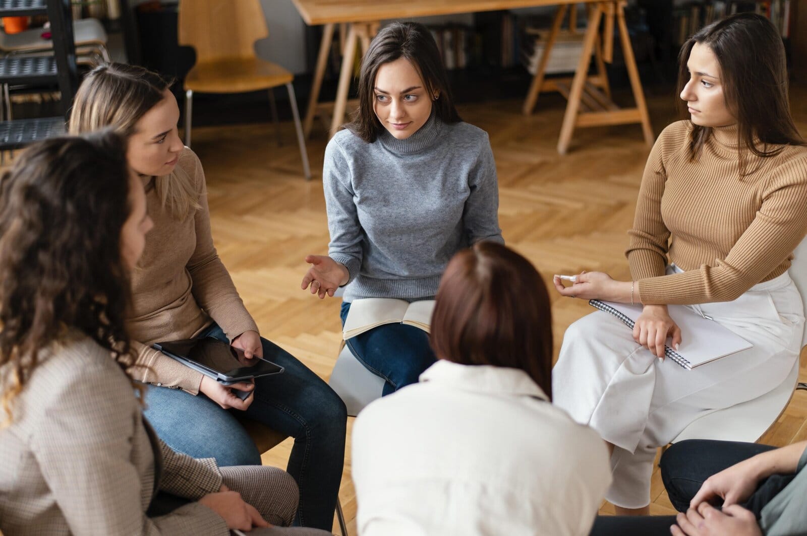 Five women sit in a circle indoors, engaged in a serious group discussion. One woman in the center gestures as she speaks, while the others listen attentively, holding notebooks and a tablet.