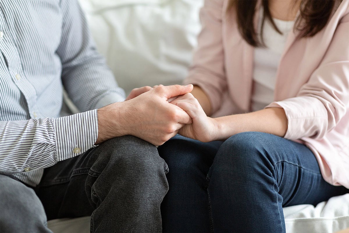 Two people sitting close together on a couch, holding hands in a comforting gesture. Only their torsos and hands are visible, suggesting a moment of support or empathy.