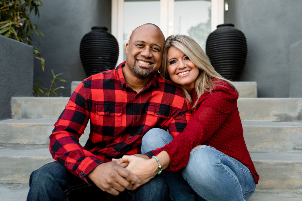 A smiling couple sits close together on outdoor steps, holding hands. The man wears a red and black plaid shirt; the woman has long blonde hair and a red blouse. Two large black vases are placed behind them.