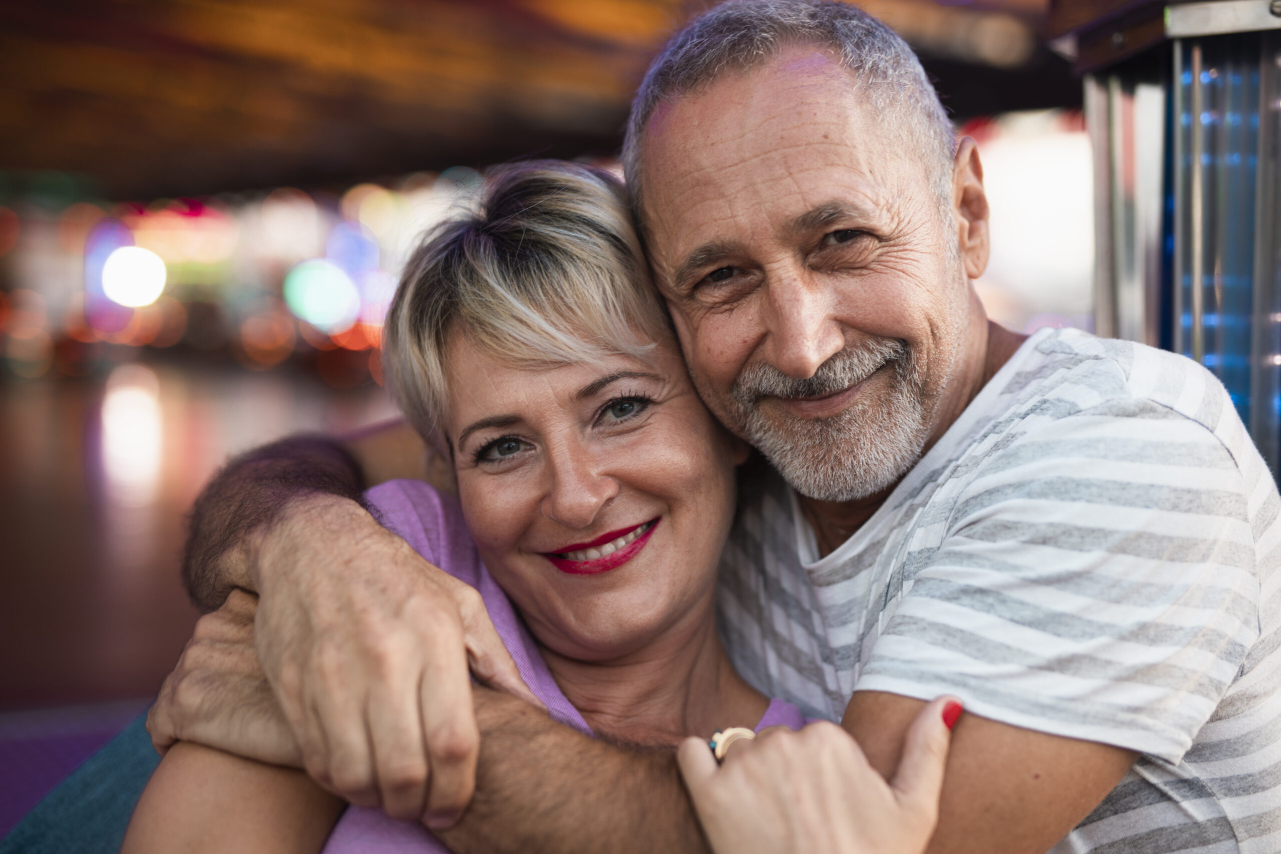 An older couple smiles warmly, embracing each other closely. The man wears a striped shirt, and the woman has short blonde hair and wears a pink top. The background is colorful and softly blurred.