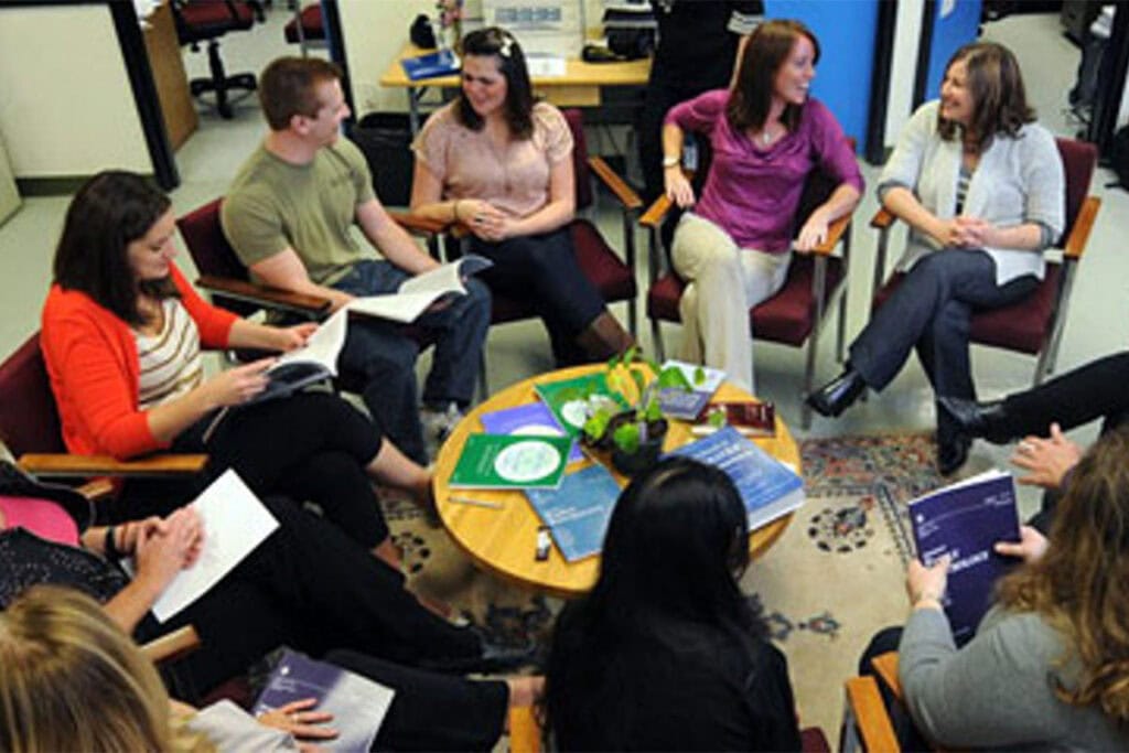 A group of people sit in a circle in an office setting, holding books and notebooks, engaged in a lively discussion around a coffee table with papers and a small plant.