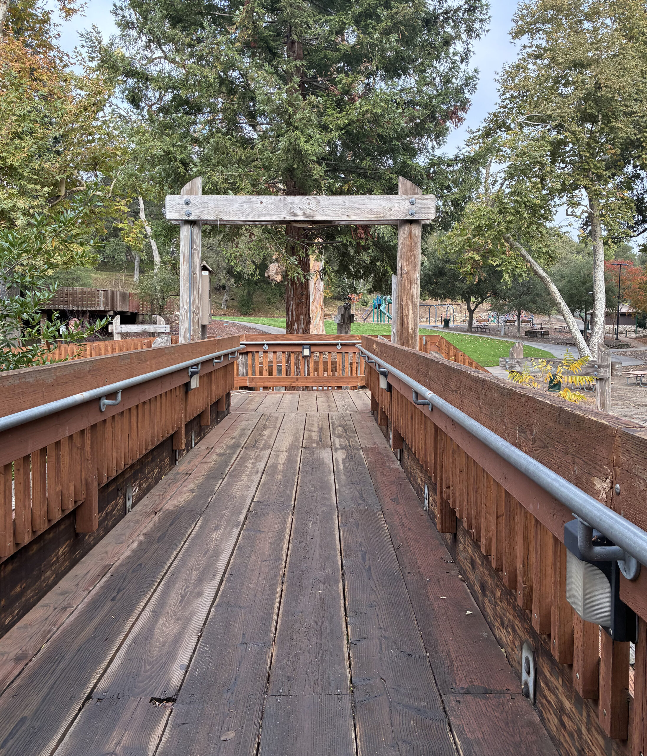 A wooden bridge with railings leads to a park area with tall trees, a playground, and benches. The scene features autumn colors and a large redwood tree in the background.