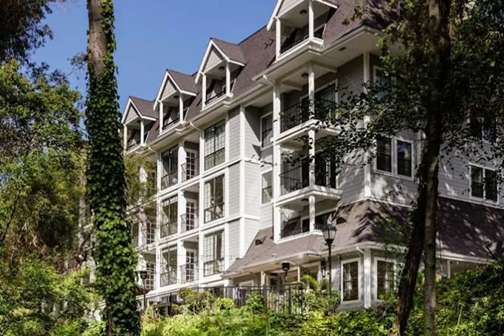 A white, four-story building with multiple balconies and dormer windows is surrounded by tall trees and greenery under a clear blue sky.
