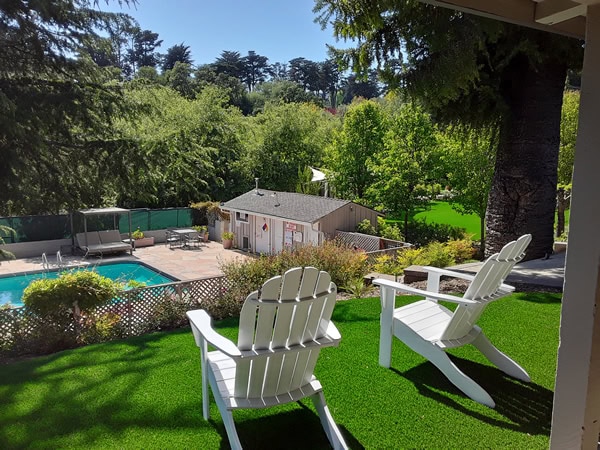 Two white Adirondack chairs sit on artificial grass overlooking a pool, patio area, and a small building, surrounded by lush trees and greenery under a clear sky.
