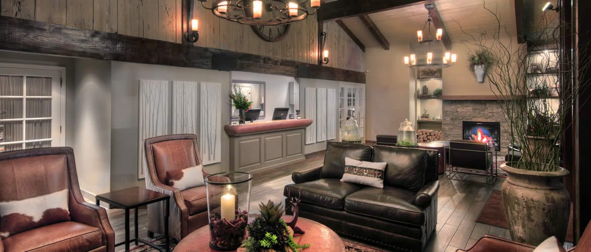 A cozy, modern lobby with leather seating, a lit fireplace, rustic wooden beams, soft lighting, and a reception desk in the background. Decorative plants and candles add warmth to the inviting space.
